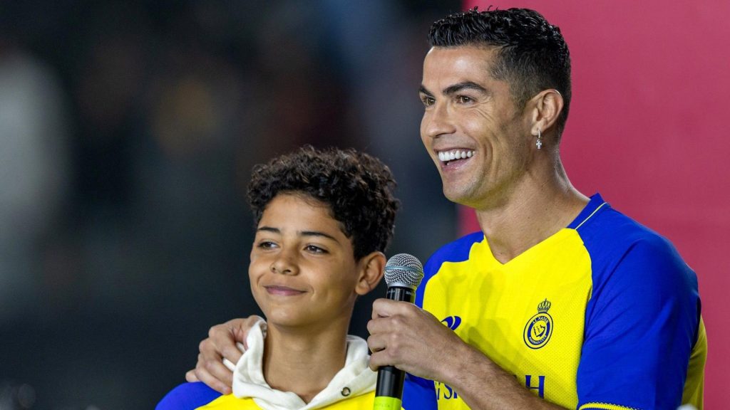 Cristiano Ronaldo and his son Junior at Al Nassr smiling while Cristiano Ronaldo is holding a microphone