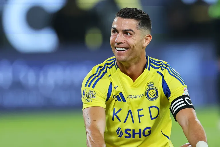 Frontview of Cristiano Ronaldo in na Al Nassr shirt sitting on the pitch and smiling with his mouth open, while facing left and holding his knees